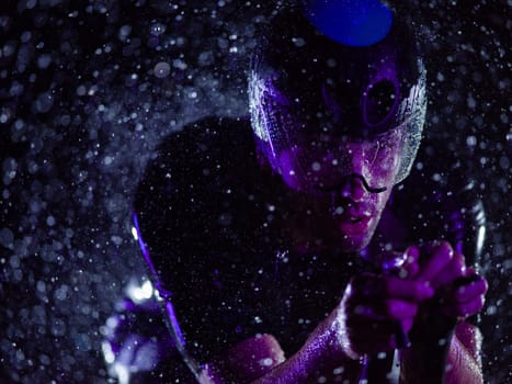 A triathlete braving the rain as he cycles through the night, preparing himself for the upcoming marathon. The blurred raindrops in the foreground and the dark, moody atmosphere in the background add to the sense of determination and grit shown by the athlete