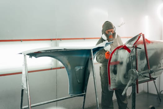 Auto mechanic worker wearing protective workwear spraying white paint on car part at workshop close up