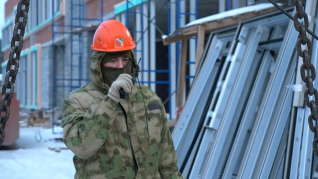 Man in helmet commands into walkie-talkie at construction site. Clip. Man talks on walkie-talkie at construction site. Contractor at construction site with walkie-talkie.