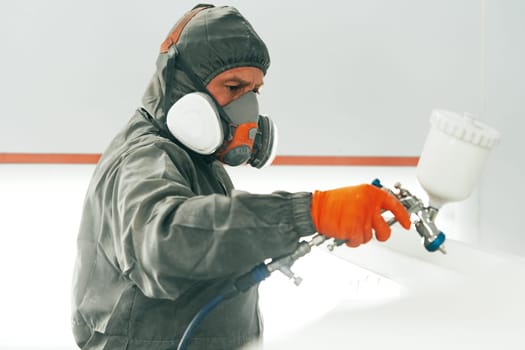 Auto mechanic worker wearing protective workwear spraying white paint on car part at workshop close up