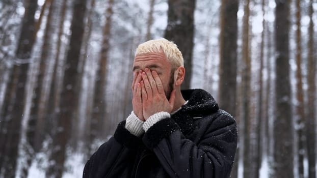 Stylish hipster man in pine tree winter forest rubbing his cold red face with his hands. Media. Portrait of a man outdoors spending time alone in winter woods
