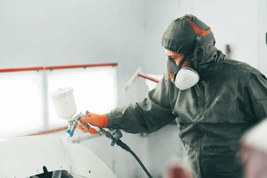 Male painter in respiratory mask and protective suit painting car with spray gun in car service close up