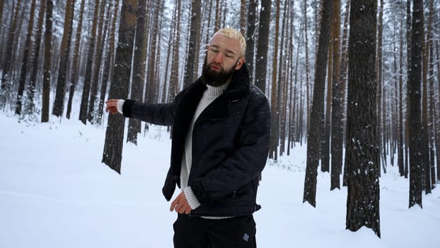 Portrait of handsome young bearded man in warm clothing standing outdoors in park on winter day. Media. Hipster blond model in woods