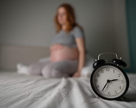 Caucasian pregnant woman sits on the bed and suffers from insomnia. Alarm clock in the foreground