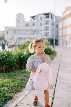 Little girl with a plush rabbit stands touching her chin with her hand near an apartment building. High quality photo