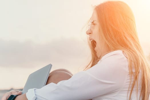 Digital nomad, woman in the hat, a business woman with a laptop sits on the rocks by the sea during sunset, makes a business transaction online from a distance. Freelance, remote work on vacation.