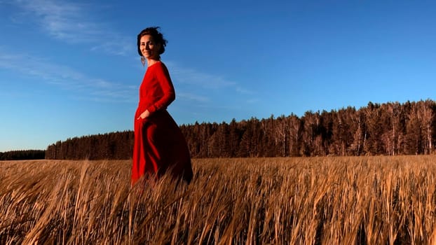 Young woman model in scarlet dress flowing in the wind. Stock clip. Brunette woman with curly hair in fild with forest and blue sky on the background