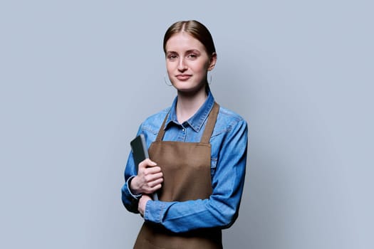 Young smiling female worker in apron uniform holding digital tablet on gray studio background. Digital technologies in business work, Internet apps applications for online orders and customer service