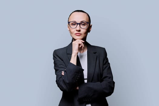 Portrait of thinking serious 30s business woman on grey studio background. Confident female in glasses, suit looking at camera. Business work teaching job career people concept