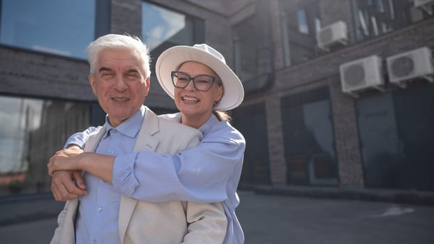 A woman in a hat and glasses hugs an elderly man in a white jacket from behind. Romantic relationships of mature people