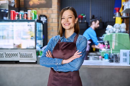 Confident successful young woman service worker in apron with crossed arms looking at camera in restaurant cafeteria coffee pastry shop. Small business, staff, occupation, entrepreneur, owner, work