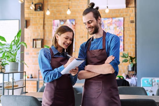 Small business team confident successful colleagues partners owners young man woman in aprons writing at working notebook at workplace in restaurant coffee shop cafeteria. Partnership teamwork work