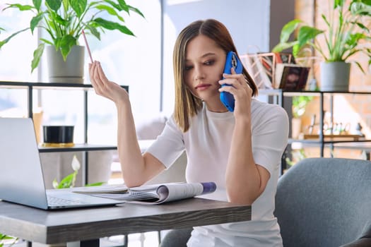 Young woman sitting at table with laptop computer talking on mobile phone in coworking cafe. Female university college student preparing for test exam, freelancer, business employee working remotely