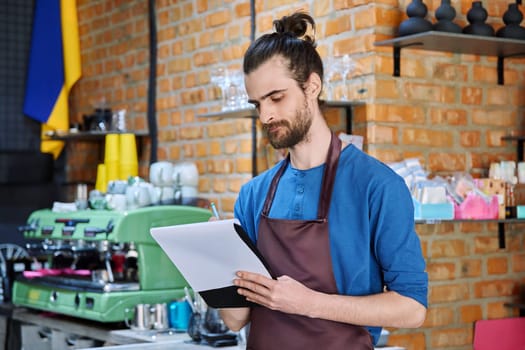 Young serious man in apron, food service worker, small business owner entrepreneur with work papers near counter of coffee shop cafe cafeteria. Staff occupation successful small business work concept