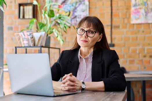 Middle-aged business confident woman working remotely at table with laptop computer looking at camera in coworking cafe. Business, mature people, success, leadership, management, empowerment concept