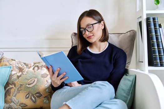 Young woman reading a book, sitting on the couch at home, engrossed in fiction. Literary hobby, education, lifestyle, youth concept