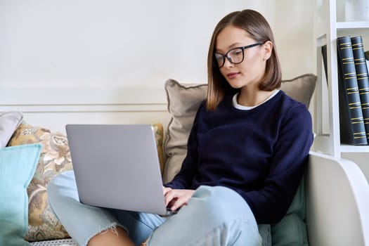 Young woman in glasses working at home, sitting on couch using laptop computer. Freelancing, training, education, remote work, leisure, internet online technology applications apps concept