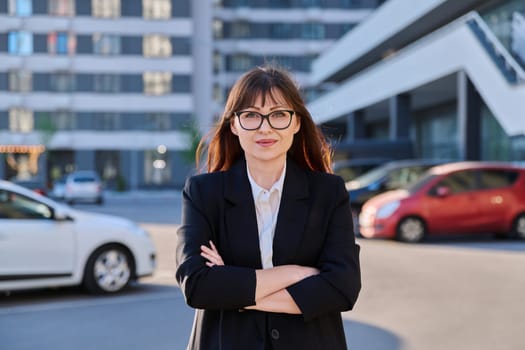 Mature confident successful business woman with crossed arms in black suit looking at camera outdoors, backdrop of modern city. Business, entrepreneurship mentoring insurance sales advertising work