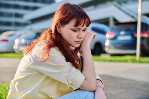 Upset sad unhappy young female sitting on steps. Pensive serious beautiful red-haired girl student 19-20 years old sitting outdoor. Problems, difficulties, depression, mental health of young people