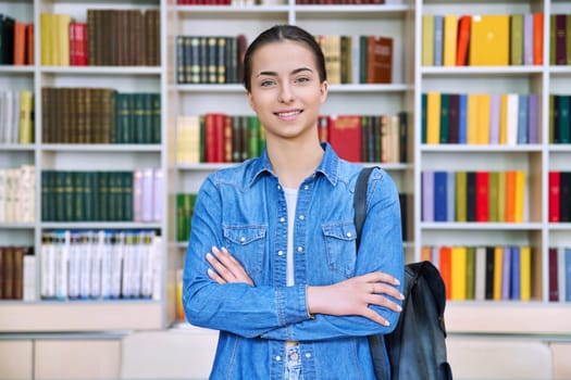 Portrait of smiling teenage girl student. Confident female teenager 16,17 years old with backpack, crossed arms inside high school building, background of library shelves books. Education, adolescence