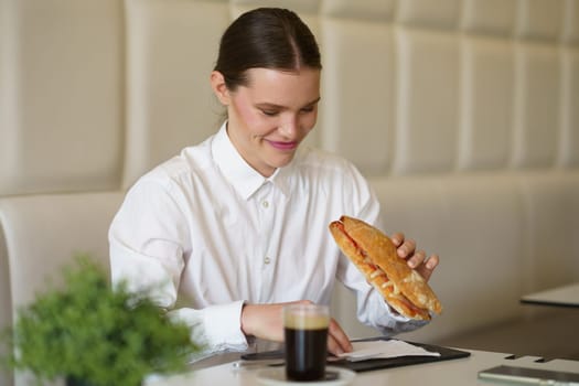 Elegant caucasian adult beauty businesswoman eating a sandwich and drinking coffee in a modern cafeteria