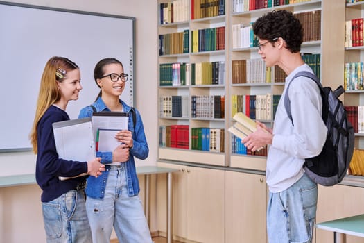 Group of teenage high school students in library classroom. Three schoolchildren 16, 17 years old, talking laughing together. Education, adolescence, friendship communication concept