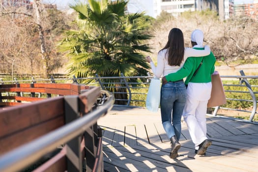 rear view of two unrecognizable young female friends walking through a city park with shopping bags, concept of friendship and urban lifestyle