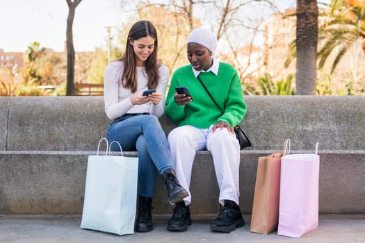 two young female friends using mobile phone sitting in a city park with shopping bags, concept of technology of communication and urban lifestyle