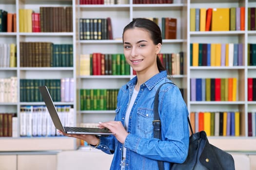 Teenage girl student with backpack using laptop computer, looking at camera inside high school building in library. Technologies, internet mobile educational apps applications, services, e-learning