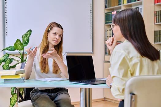 Teenage student girl studies with teacher inside classroom library. Education, high school, learning, adolescence, teaching concept