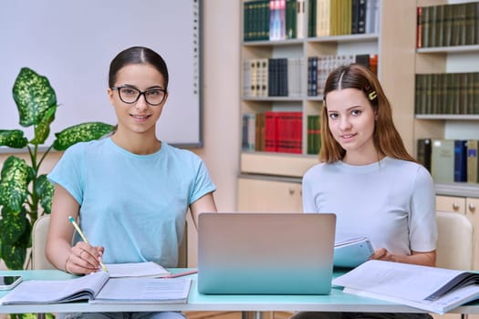 Teenage girls high school students sitting at desk in library classroom, smiling looking at camera, studying together. Education adolescence communication concept