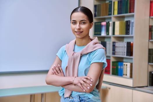 Portrait of smiling teenage girl student. Positive female teenager 16,17 years old with crossed arms inside high school building, background of library shelves books. Education, adolescence, lifestyle