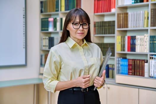 Middle aged elegant woman holding laptop computer inside library. Confident serious business female teacher professor, leader, manager, mentor, psychologist, counselor, social worker at workplace