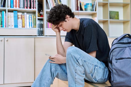 Sad upset young male college student looking at smartphone while sitting in classroom. Negative emotions, difficulties, troubles, youth concept