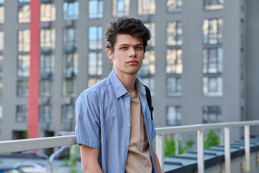 Portrait of smiling, handsome student guy 19, 20 years old, looking at camera outdoors, modern building background. Youth, urban style, young man, lifestyle concept