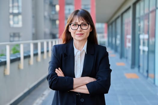 Mature confident successful business woman with crossed arms in black suit looking at camera outdoors, backdrop of modern city. Business, entrepreneurship mentoring insurance sales advertising work