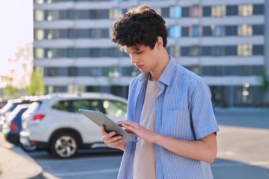 Young male using digital tablet on city street. Handsome guy student 19-20 years old, using pad for leisure, studying, working. Technology, youth, education, urban style concept