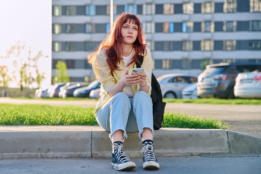 Young beautiful serious red-haired woman using smartphone sitting on floor sunny day outdoor city urban style. Youth lifestyle technology internet online applications for leisure tourism learning work