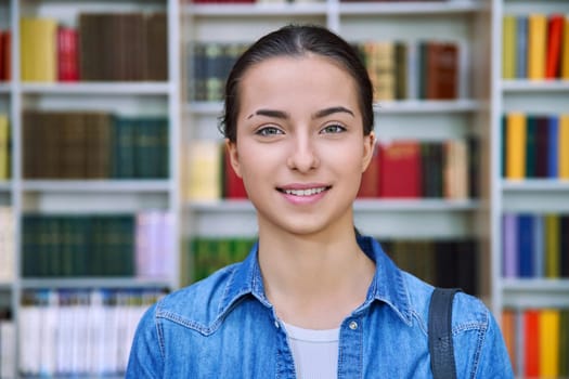 Headshot portrait of smiling teenage girl student. Confident female teenager 16,17 years old with backpack inside high school building, background of library shelves books. Education, adolescence