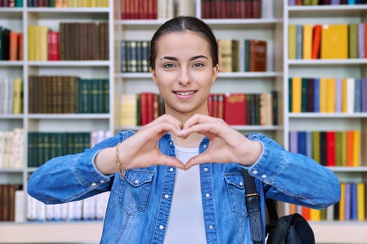 Happy teenage girl student showing heart gesture at camera with hands inside high school building. Joyful positive teenage girl 16, 17 years old in library. Emotions, education, lifestyle, adolescence