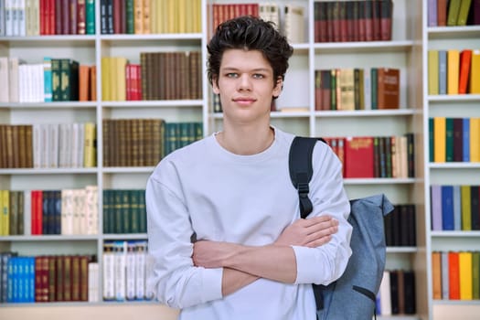 Portrait of confident handsome college student guy with crossed arms inside library of educational building. Education, youth, lifestyle concept