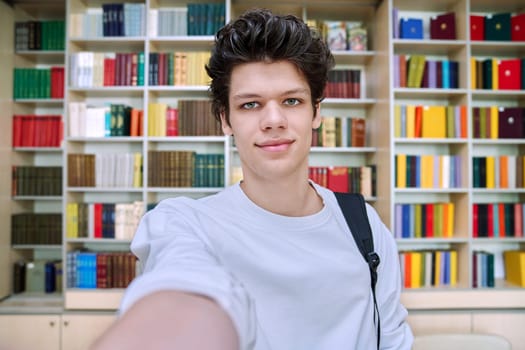 Selfie portrait of smiling handsome college student guy inside library of educational building. Education, youth, lifestyle concept