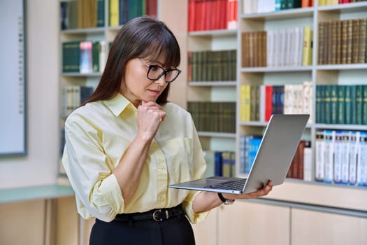 Middle aged elegant woman using laptop computer inside library. Confident serious elegant business female teacher mentor professor, leader manager, psychologist counselor, social worker at workplace