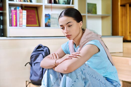 Upset sad teenage girl high school student sitting on floor in library classroom. Depression, stress, problems, loneliness, adolescence, mental health concept