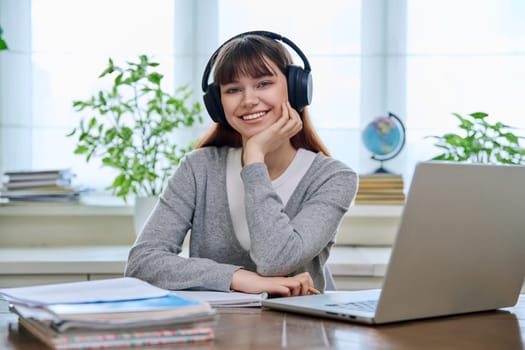 Portrait of teenage college student wearing headphones, sitting at desk, smiling and looking at camera. Education, training, youth 18,19, 20 years old