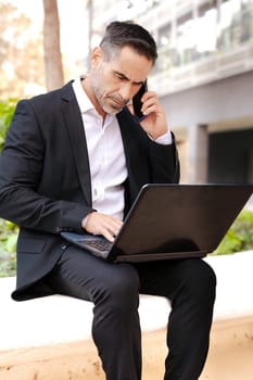 Middle Aged Businessman Dressed in Formal Attire Having A Call With Phone, Working With Laptop