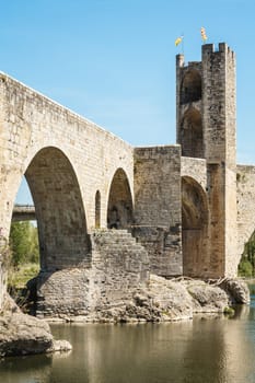 The bridge of the medieval village of Besalú with a tower in the background. The bridge is made of stone and has a stone arch. The water is calm and the sky is clear.