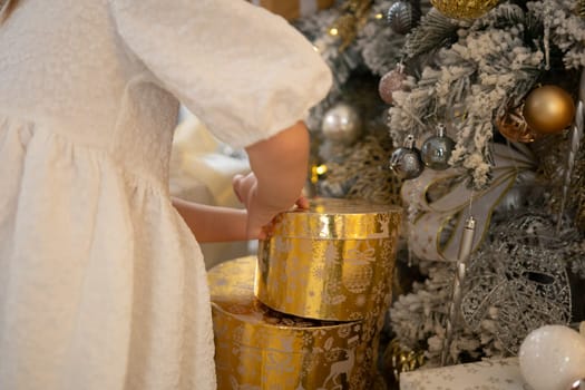 A woman is opening a gold box with a silver ball inside. The box is decorated with gold and silver ornaments, and the woman is wearing a white dress. The scene is set in a room with a Christmas tree