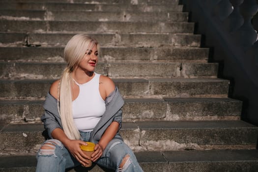 A blonde woman sits on a set of stairs, holding a cup. She is wearing a gray jacket and blue jeans