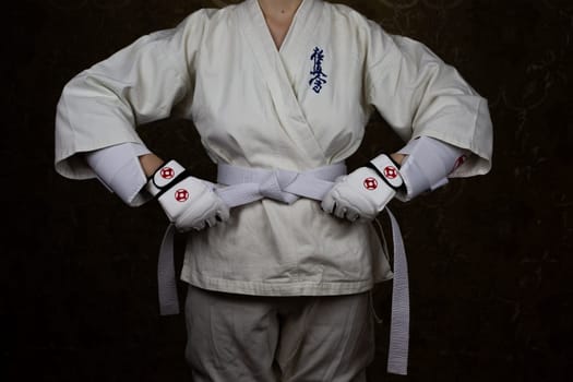 Girl Kyokushin karate athlete in kimono and protective gloves tightens white belt around her waist. Inscription in Japanese: Kyokushinkai